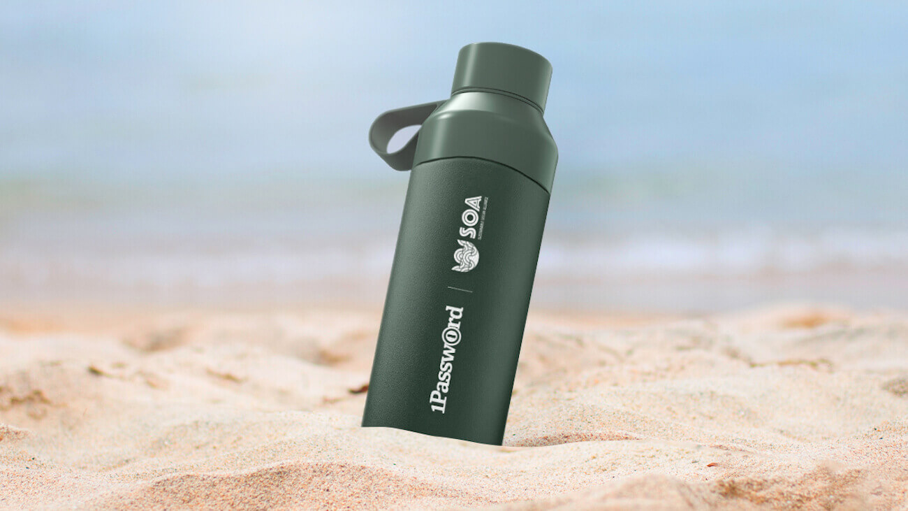 A green reuseable water bottle with 1Password and Ocean Alliance logos on it resting on the sand at the beach.